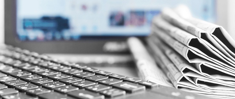 Banner Image of a View Across a Desktop Showing Computer Screen, Keyboard and a Stacked Pile of Newspapers